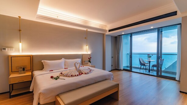 Una habitación de hotel moderna con una cama grande, mesitas de noche, un banco y un balcón con vista al mar.