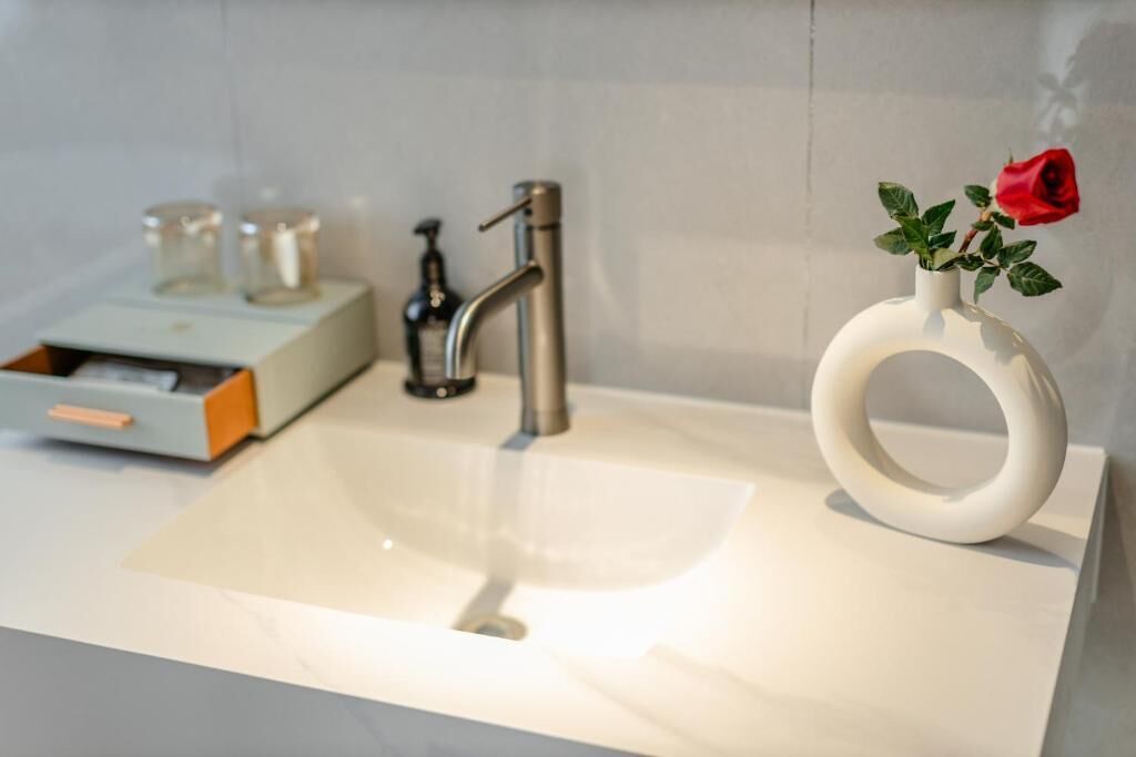 A bathroom with a white sink, a modern faucet, a glass soap dispenser, and a white vase with a red rose.