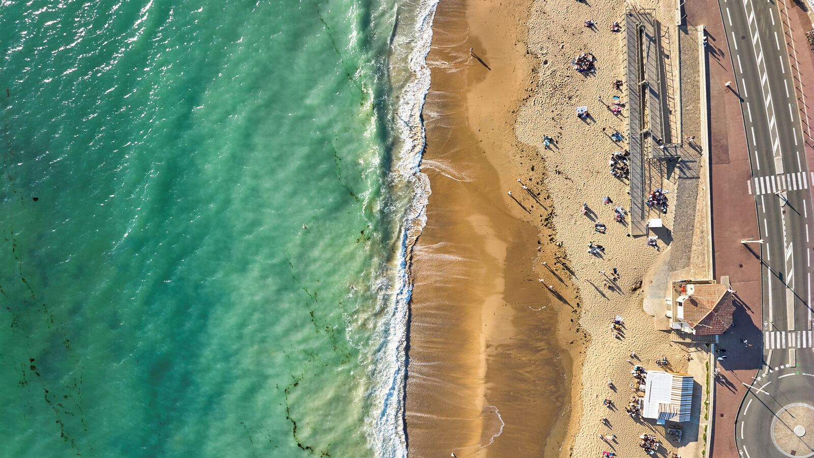 Une vue aérienne d’une plage, avec une route parallèle au rivage, quelques bâtiments et des personnes dispersées le long de la plage.
