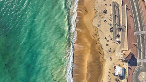 Une vue aérienne d’une plage, avec une route parallèle au rivage, quelques bâtiments et des personnes dispersées le long de la plage.