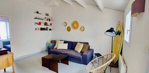 Ein Wohnzimmer mit einer Couch, einem Couchtisch und einem Sessel. Regale mit diversen Gegenständen und ein Surfbrett an der Wand.