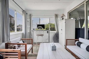 A sunlit room with a white sofa, wooden chairs, a table, and a TV on a stand. - Private Beach -Single-Level Cottage  | Sleeps 6 (East Lyme)