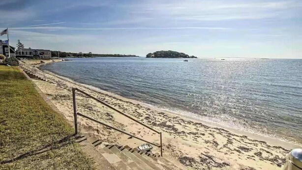 A beach with a staircase leading down to the water, a small island in the distance, and a building on the left.