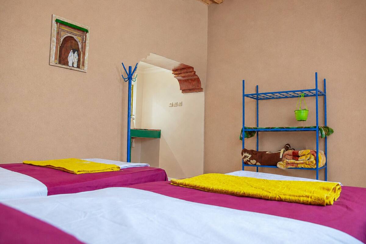 Habitación con dos camas, una litera, estante en la pared con un cubo y una manta, y una puerta con arco decorativo.