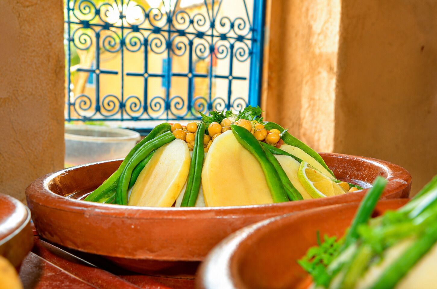A terracotta dish with sliced vegetables and chickpeas, placed on a table with a view of a blue gate.