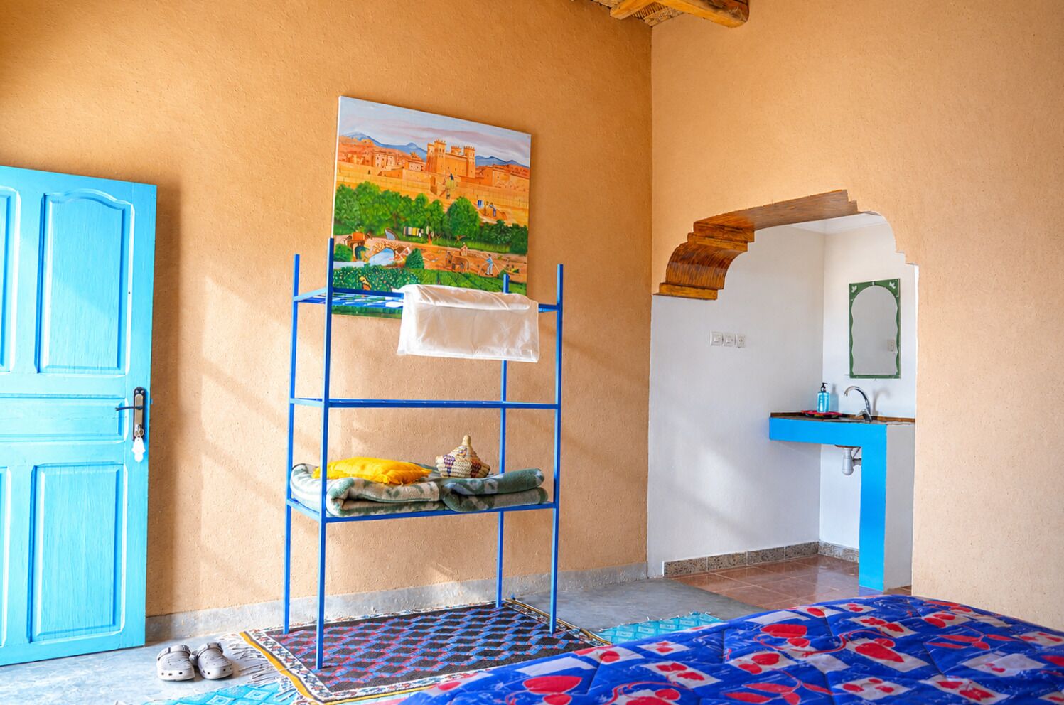 Een kamer met een blauwe deur, een wandplank met een schilderij, een wastafel met een blauw aanrecht en een kamerbreed tapijt met een patroon.