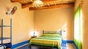 Una habitación con una cama, una escalera metálica azul, una pequeña mesa con un farol y una alfombra estampada.