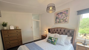 A bedroom with a bed, dresser, nightstand, lamp, window, and artwork.