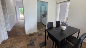 A room with a dining table, chairs, a potted plant, and a kitchen area visible through an open door.