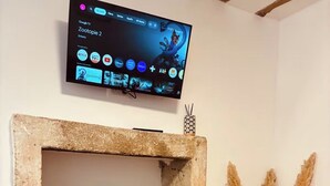 Een kamer met een stenen open haard, een houten dressoir, een ronde houten tafel en een flatscreen televisie die boven de open haard is bevestigd, waarop een kinderprogramma wordt vertoond.