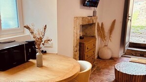 Een kamer met een ronde houten tafel, twee stoelen, een aan de muur bevestigde televisie, een stenen vloer en een raam met uitzicht.