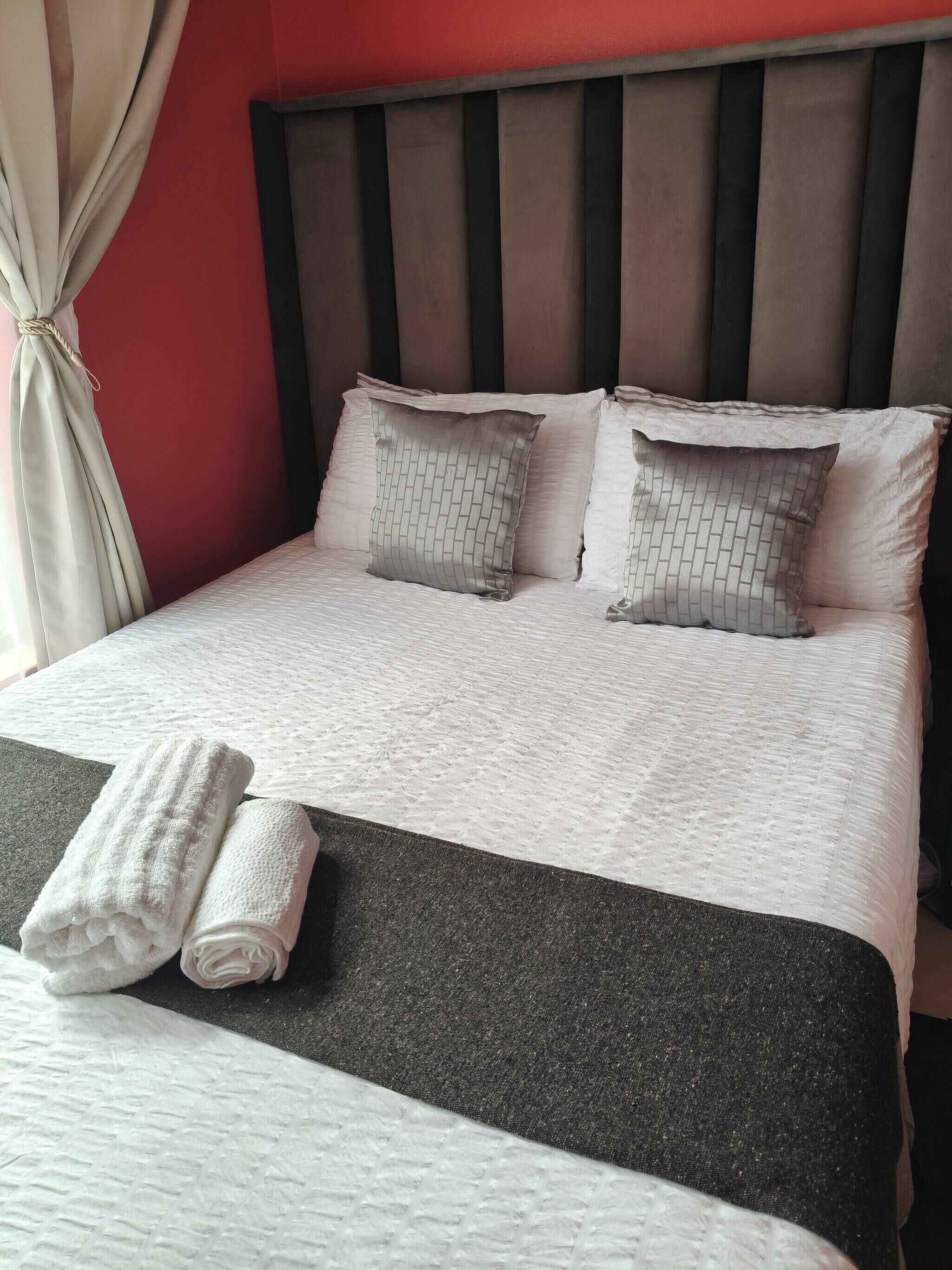 Una cama con colcha blanca y gris, dos almohadas y dos toallas enrolladas.