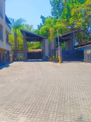A paved courtyard with a covered entrance, buildings on either side, and trees in the background.