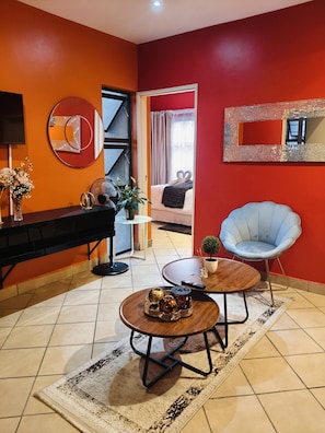 A modern living room with a sofa, a coffee table, a blue chair, a TV, and a dining area with a table and chairs.