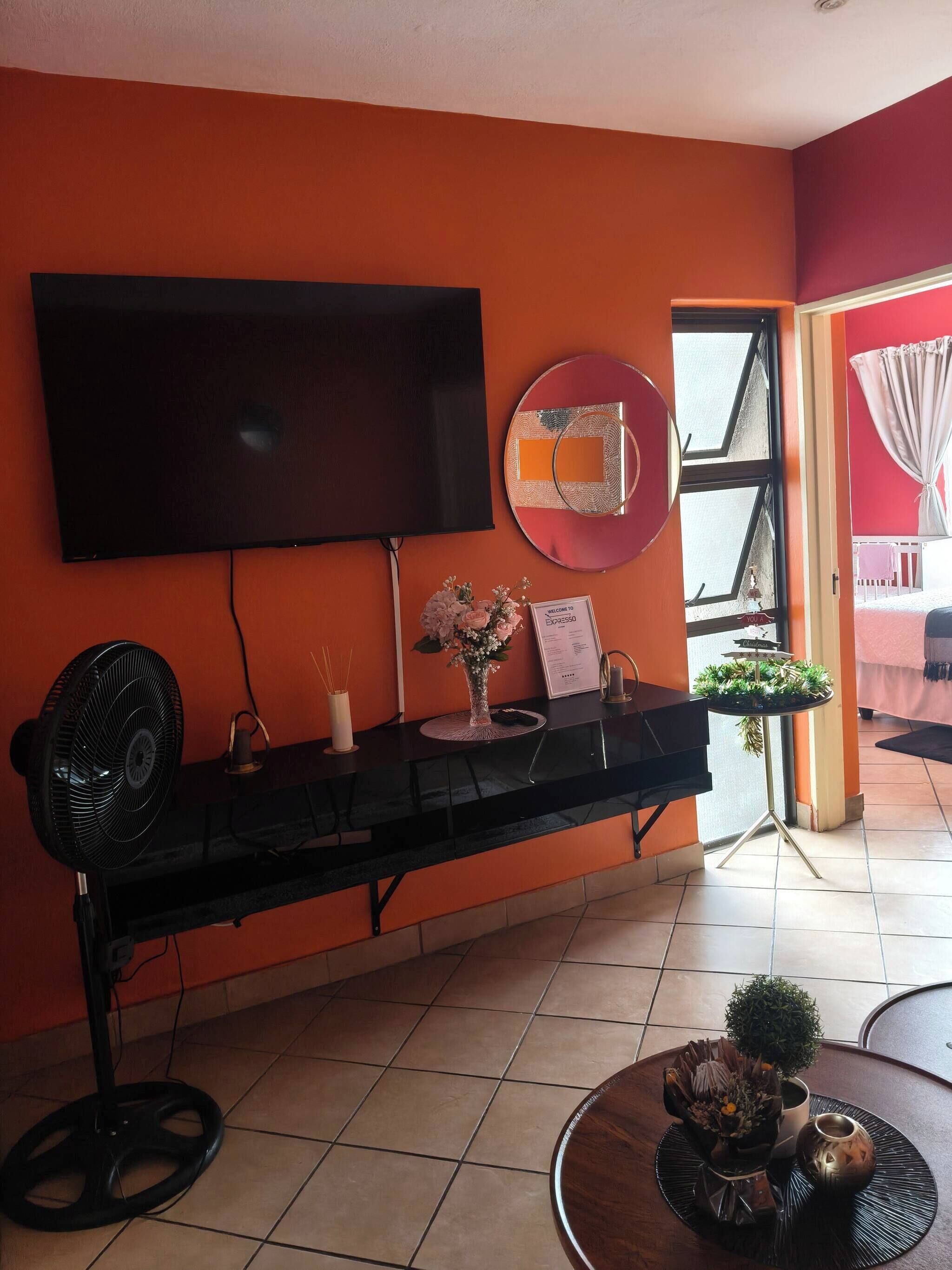 Habitación con mostrador de recepción negro, televisor de pantalla plana, ventilador, jarra con flores y una mesita con planta y vela.