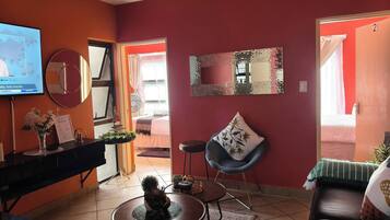 Una sala de estar con televisor, una mesa redonda de centro, un sofá de cuero negro, un espejo y una puerta que da a otra habitación.