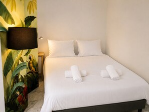 A hotel room with a bed, white linens, two rolled towels, a nightstand, a lamp, and a wall mural of parrots. - 16672352 LIONS 1-Bedroom LOUIS BLANC: Elegance and Light (Cannes)