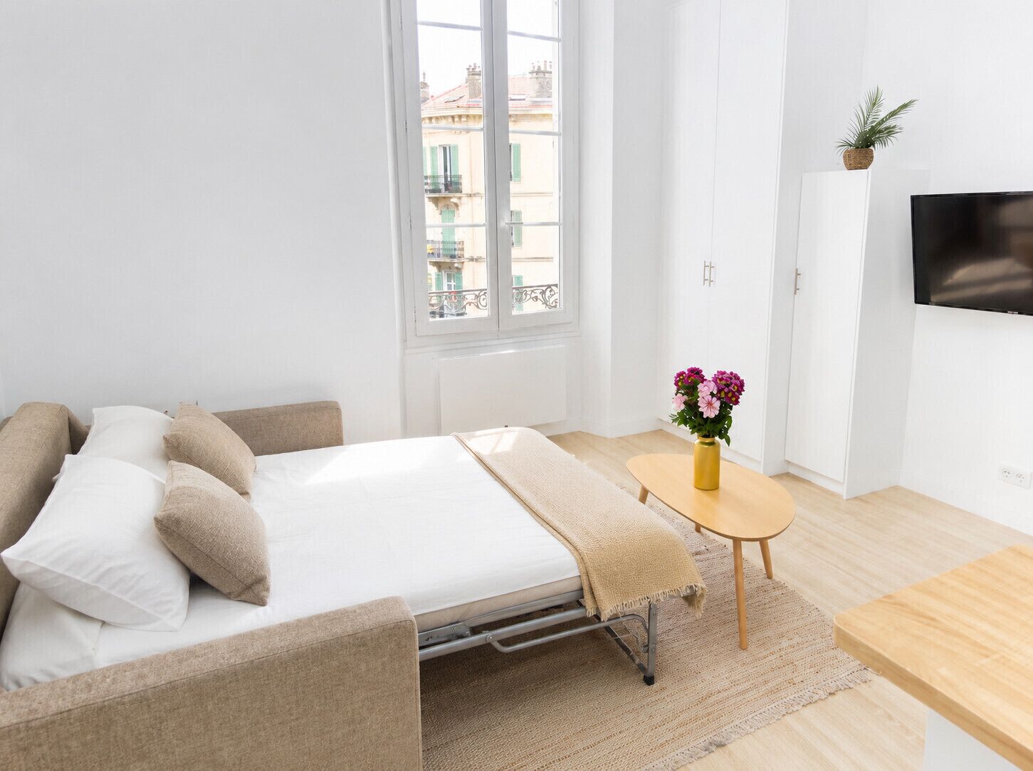 A modern bedroom with a bed, a small table with flowers, a TV, and a view of buildings through a window.