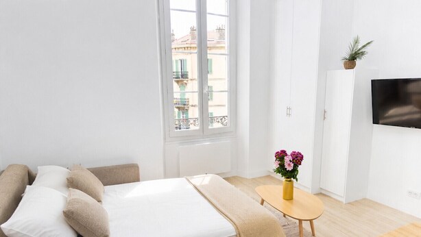 A modern bedroom with a bed, a small table with flowers, a TV, and a view of buildings through a window. - 16664494LIONS Renovated studio, unobstructed and (Cannes)