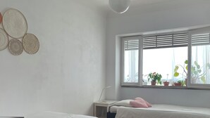 A bedroom with two beds, a window with blinds, a bedside table, and a hanging light fixture. - Bright and spacious, just 20 meters from the subway station in a family-friendly neighborhood. (Lisboa)