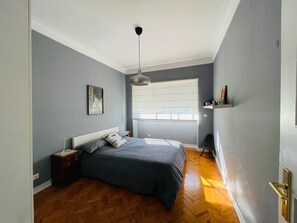 A bedroom with a bed, a nightstand, a wall-mounted shelf, a window with blinds, and a hanging light fixture. - Bright and spacious, just 20 meters from the subway station in a family-friendly neighborhood. (Lisboa)