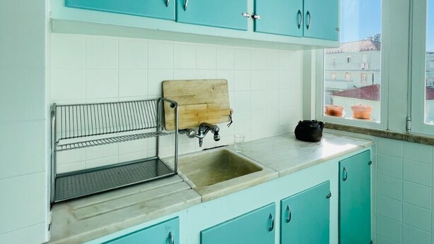 A kitchen with teal cabinets, a wooden cutting board, a metal rack, and a window with a view. - Bright and spacious, just 20 meters from the subway station in a family-friendly neighborhood. (Lisboa)