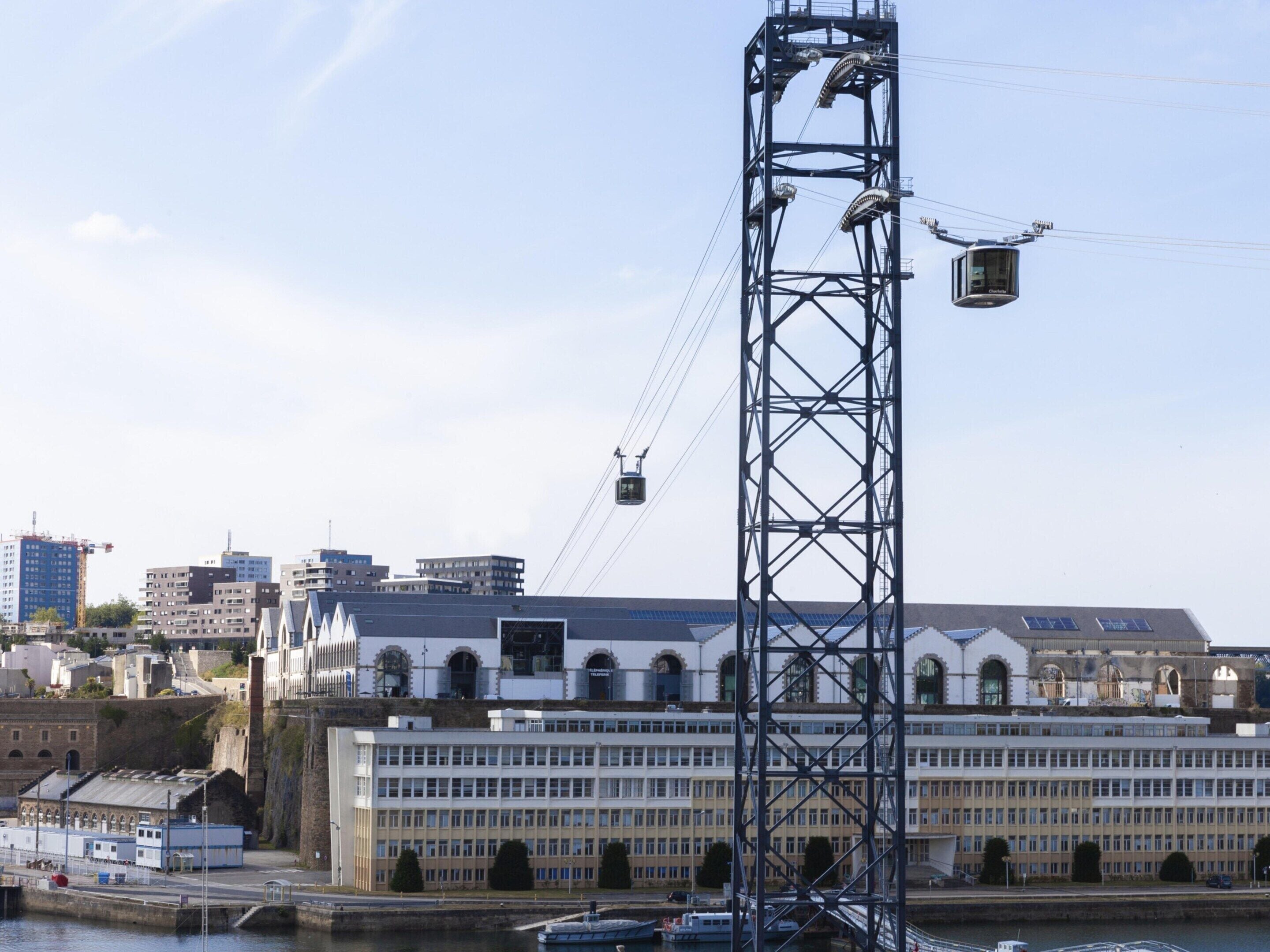 Una estructura metàl·lica alta amb un sistema de telefèric, un edifici gran amb finestres d'arc i una massa d'aigua amb vaixells.