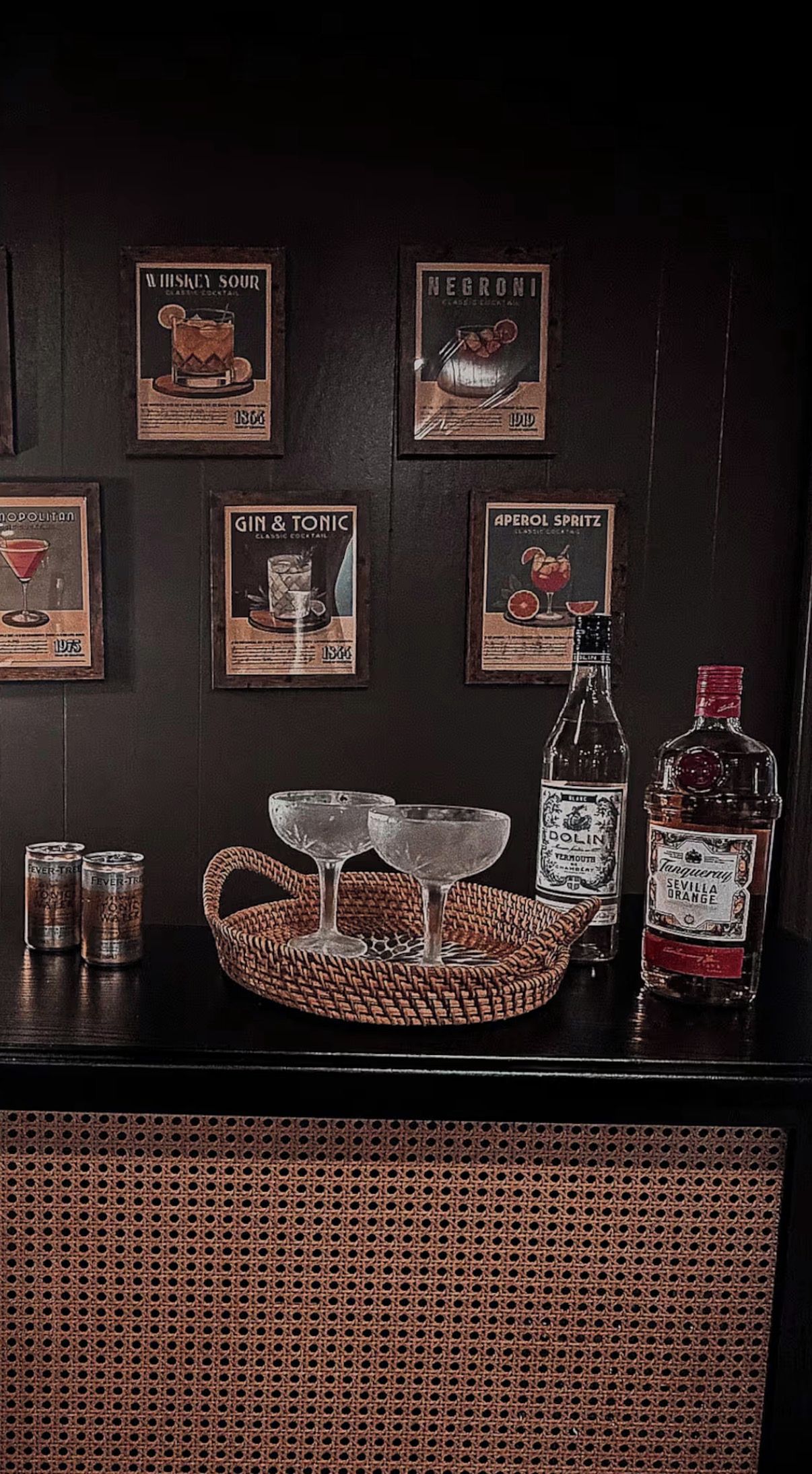 A bar area with framed cocktail posters, a bottle of DOLIN Vermouth, a bottle of Tanguraw Orange, and a woven basket with glasses.