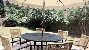 An outdoor patio with a round table and six wicker chairs, shaded by a large umbrella. - The Long Weekend Cottage Charming English Cottage Minutes from Main Street (Fredericksburg)