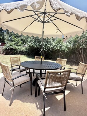 Eine Terrasse im Freien mit einem runden Tisch und sechs Korbstühlen, beschattet von einem großen Sonnenschirm.