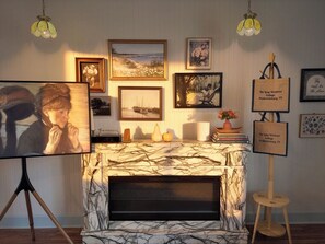 A room with a fireplace, framed artwork on the wall, and a small table with decorative items. - The Long Weekend Cottage · 2 Minutes from Main Street & 8 Minutes from Wineries (Fredericksburg)