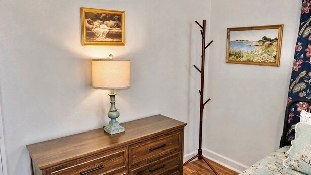 A bedroom with a wooden dresser, a lamp, two framed pictures on the wall, and a floral-patterned bedspread. - The Long Weekend Cottage Charming English Cottage Minutes from Main Street (Fredericksburg)
