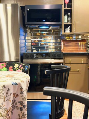 A compact kitchen with a microwave, oven, and a wooden cutting board with utensils. - The Long Weekend Cottage · 2 Minutes from Main Street & 8 Minutes from Wineries (Fredericksburg)