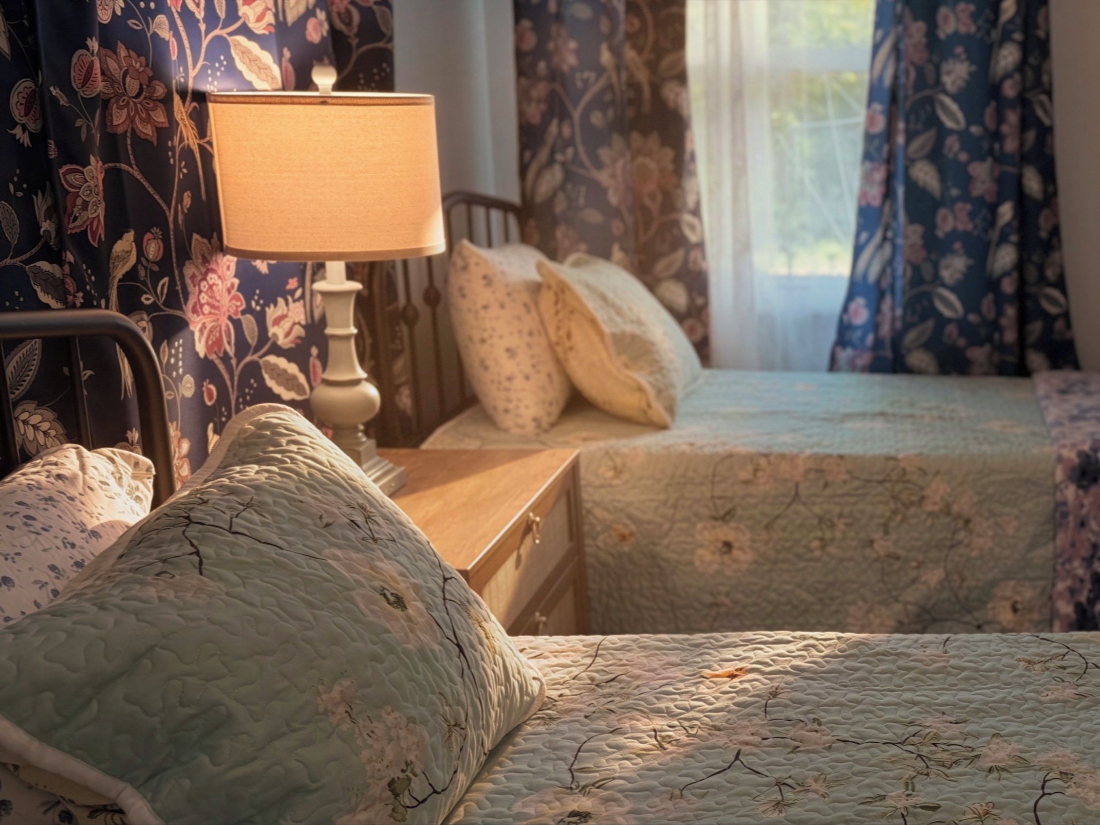 A bedroom with two beds, a nightstand, a lamp, and floral curtains.