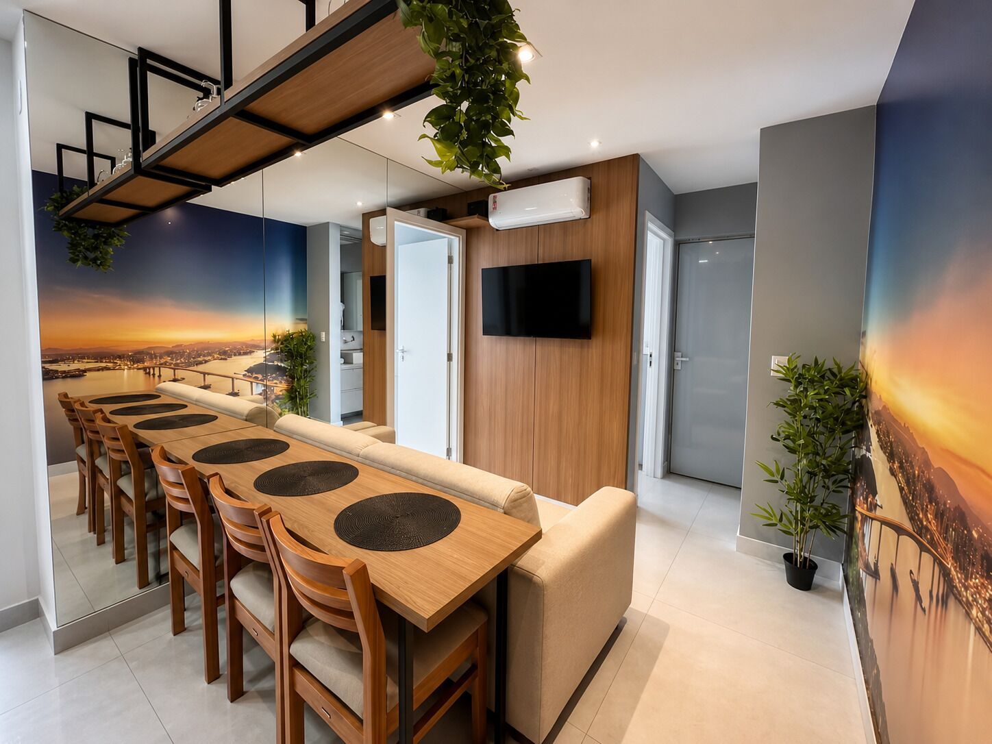 Un espace repas moderne avec une longue table en bois, plusieurs chaises et un canapé. Un téléviseur est fixé au mur et une plante est suspendue au plafond.