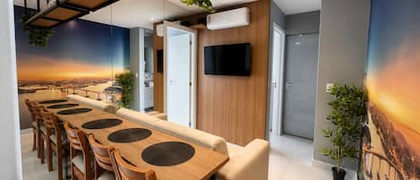 A modern dining area with a long wooden table, multiple chairs, and a sofa. There is a television mounted on the wall and a plant on the ceiling.