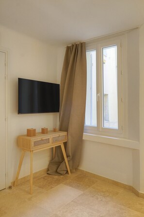 A room with a flat-screen TV mounted on the wall, a wooden console table with storage, and a window with curtains. - Townhouse Studio 2 Deluxe (Cannes)