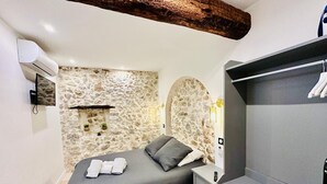 A bedroom with a stone wall, a bed with pillows, a bedside table, a wardrobe, and a mounted TV. - 1539891003 TOWNHOUSE STUDIO 2 (Cannes)