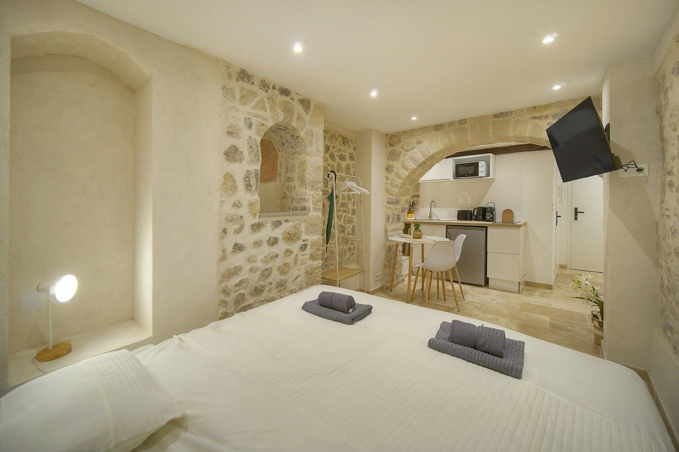 Un dormitorio con pared de piedra, una cama con ropa de cama blanca, un escritorio con silla y una kitchenette al fondo.