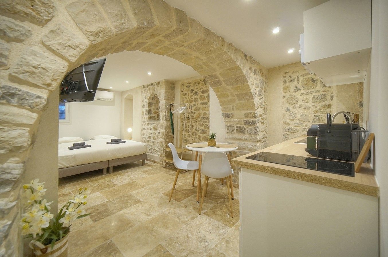 A bedroom with a bed, a small table, and chairs, a kitchenette with a stove, and a stone archway.