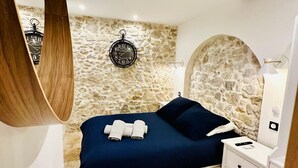 Un dormitorio con pared de piedra, cama con colcha azul oscuro, viga de techo de madera, reloj de pared redondo y mesita de noche con lámpara.