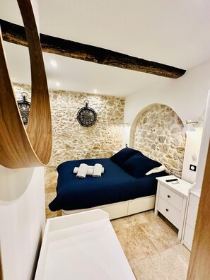 Een slaapkamer met een stenen muur, een bed met donkerblauwe dekbedovertrek, een houten balk aan het plafond, een ronde wandklok en een nachtkastje met een lamp.