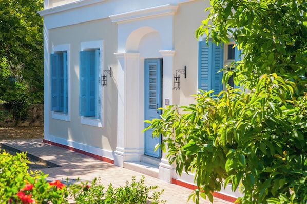 A white building with blue shutters and a red trim, surrounded by greenery. - Beachfront, Pool, Chic- Live in Style: Pyrgo Villa (Ialisos)