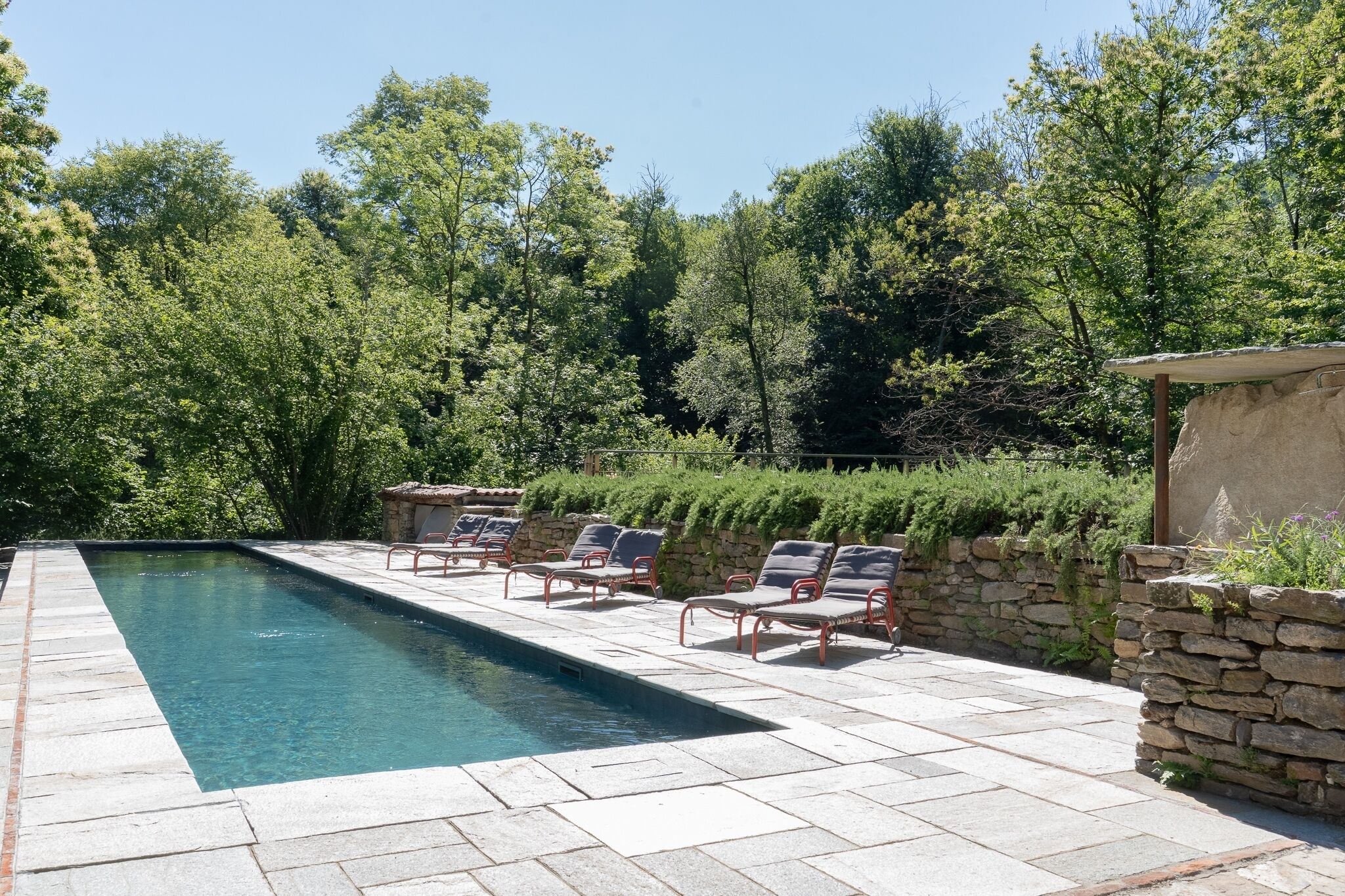 Une piscine rectangulaire bordée de pierres, entourée d’un mur de pierres et d’une rangée de chaises en métal rouge.