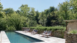 Una piscina rectangular con borde de piedra, rodeada por un muro de piedra y una fila de sillas reclinables de metal rojas.