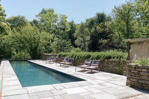 Una piscina rectangular con borde de piedra, rodeada por un muro de piedra y una fila de sillas reclinables de metal rojas.
