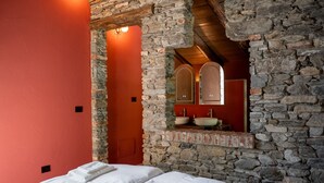 Una habitación con pared de piedra, una cama con ropa blanca y un baño con lavamanos y espejo de piedra.