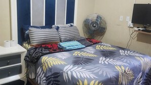 A bedroom with a bed, pillows, a bedside table with a lamp, a wall-mounted TV, and a standing fan. - 92 HOWARD GUEST HOUSE BENONI (Benoni)