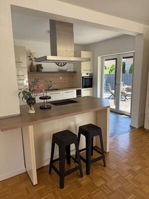 A modern kitchen with a wooden countertop, two black stools, a built-in oven, and a sliding glass door leading to an outdoor area. - Carolingian Stay (Emmerich am Rhein)