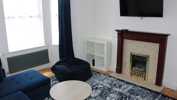 A living room with a fireplace, a flat-screen TV, a blue sofa, a round coffee table, and a large window with curtains.
